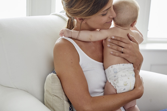 A smiling mother sits in a chair holding a diapered baby close to her chest