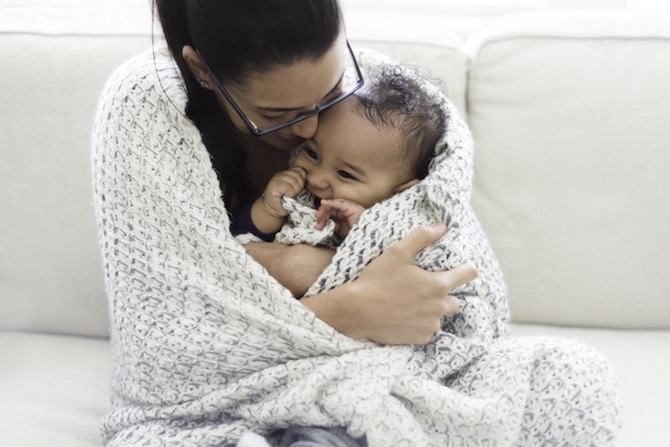 A smiling mother and baby sit on a couch wrapped in a blanket