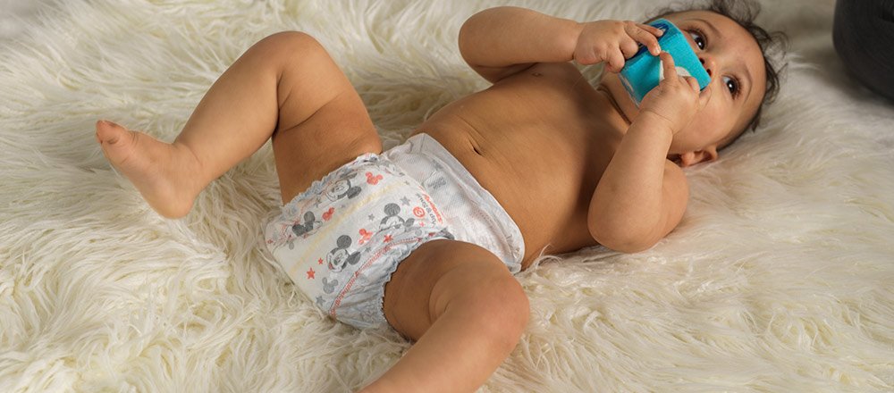 A diapered baby lays on a fuzzy blanket while chewing on a soft toy