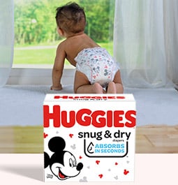 Un bebé en pañales mirando a través de una ventana en un día soleado detrás de una imagen de una caja de pañales Huggies Snug and Dry