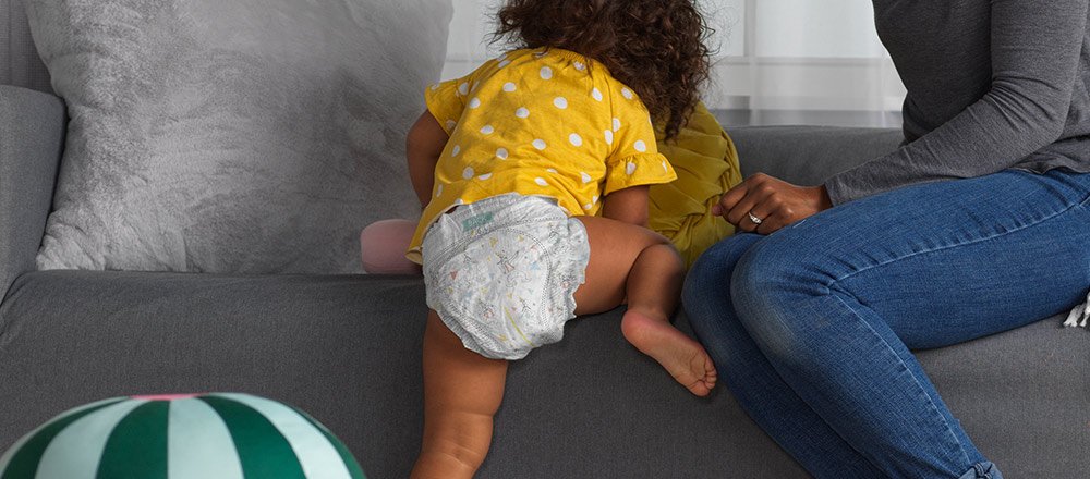 A baby wearing a Huggies Little Movers Plus Diaper climbs onto a couch next to their mother