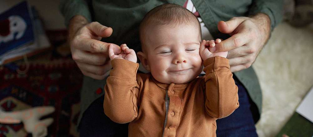 A smiling baby holds onto their father's index fingers