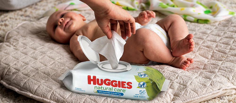 A smiling baby lays on a blanket while a hand pulls a wipe from a package of Huggies Natural Care Refreshing Wipes with EZ Pull lid